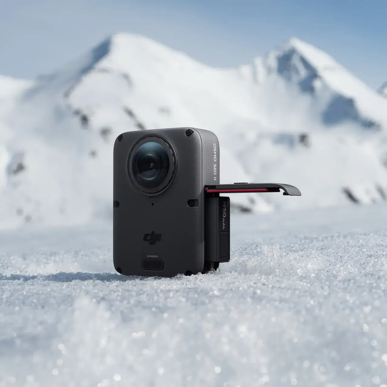 DJI OSMO 360 II 2 MARK 2, A black DJI action camera with an extended battery or accessory module, standing in snow with blurred snowy mountains in the background.